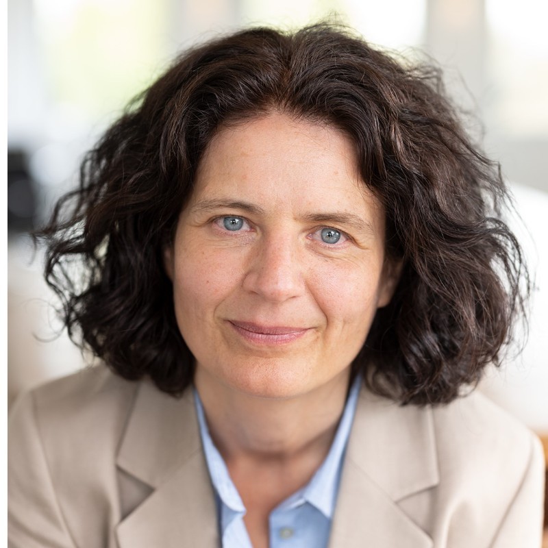 Portret van Hilde Veelaert in een beige blazer met blauwe blouse, met een neutrale achtergrond.