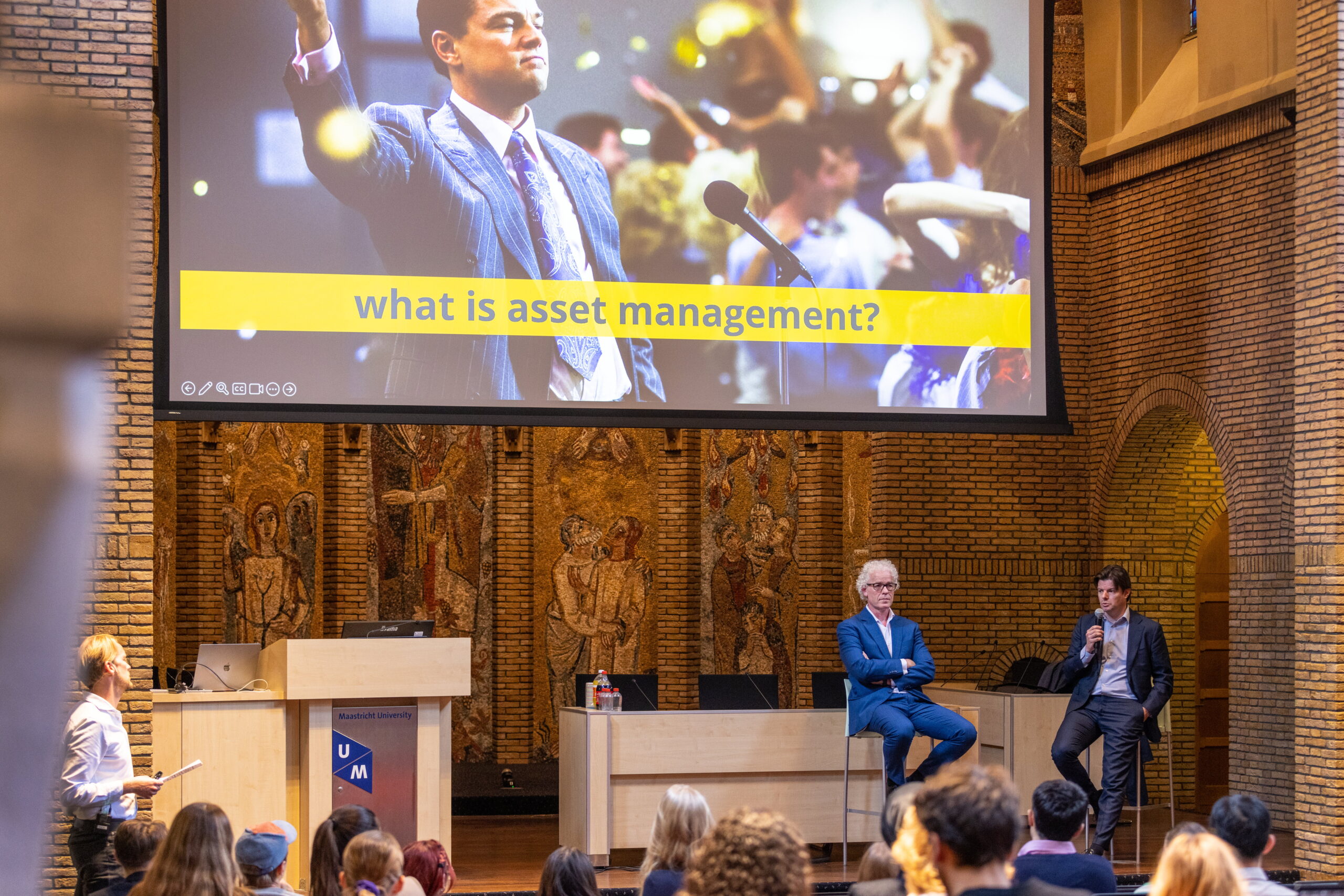 Twee sprekers beantwoorden vragen van studenten tijdens een College Tour over asset management aan de Universiteit Maastricht, met een grote projectie op de achtergrond.