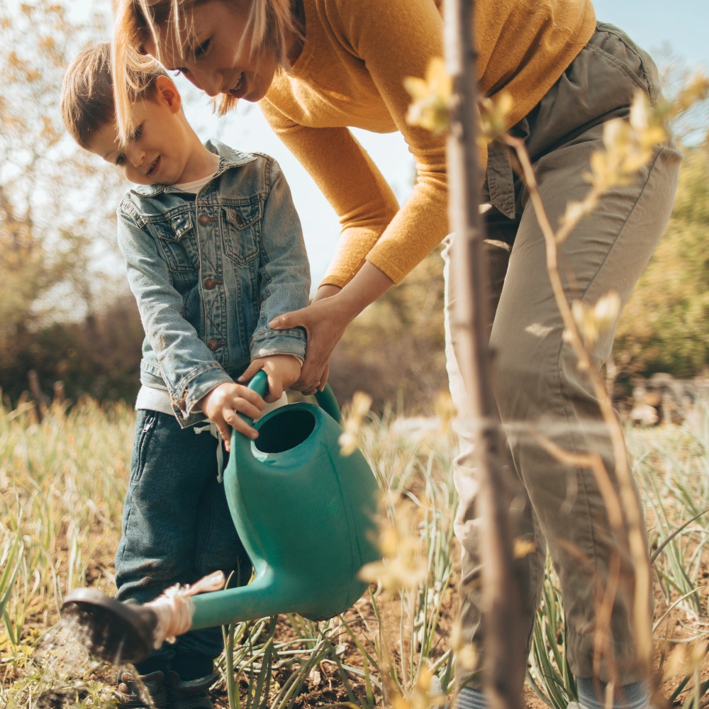 Moeder en zoon geven samen water aan een plant met een gieter in een tuin.