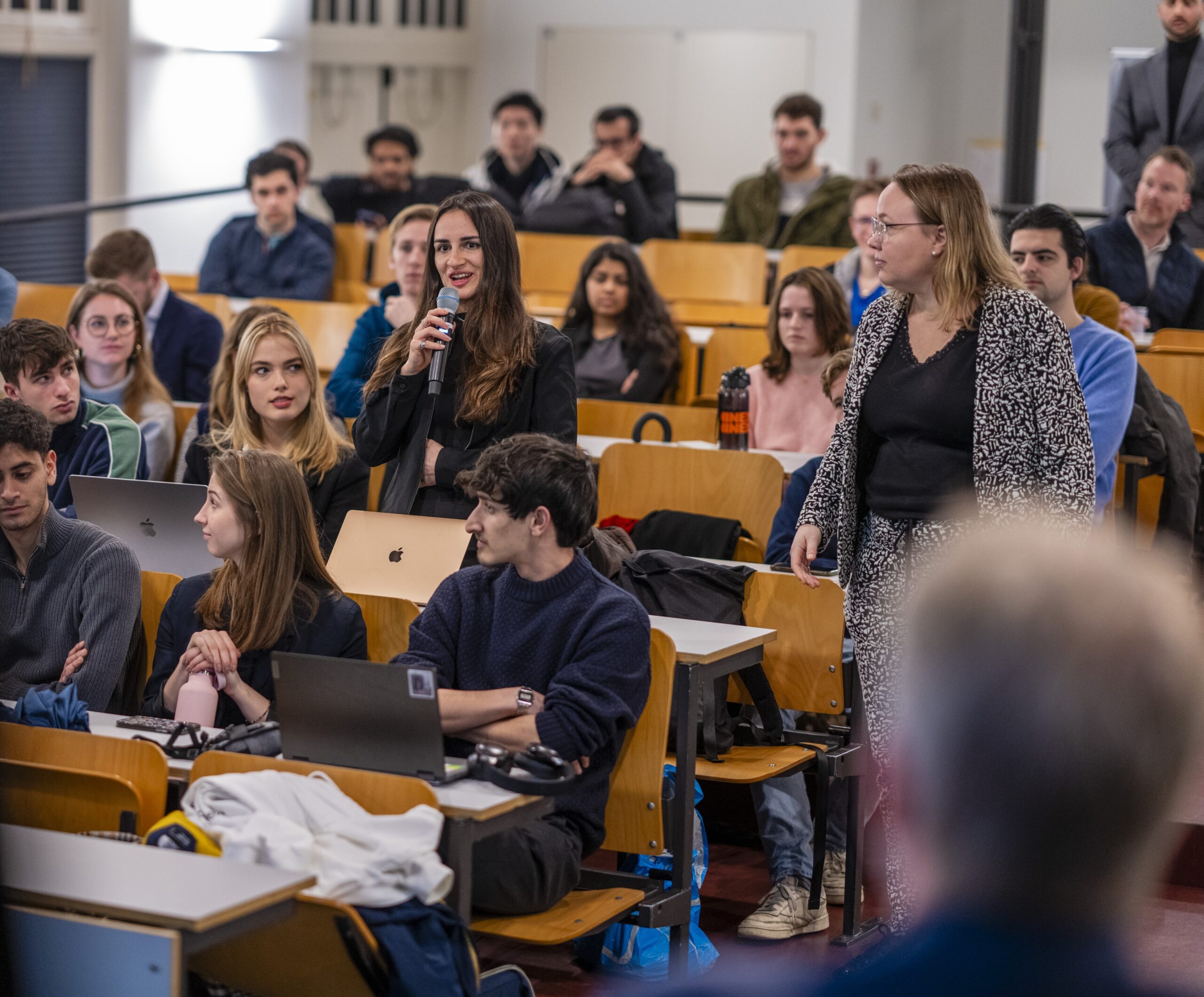 Student stelt een vraag met een microfoon tijdens een college in een volle collegezaal.