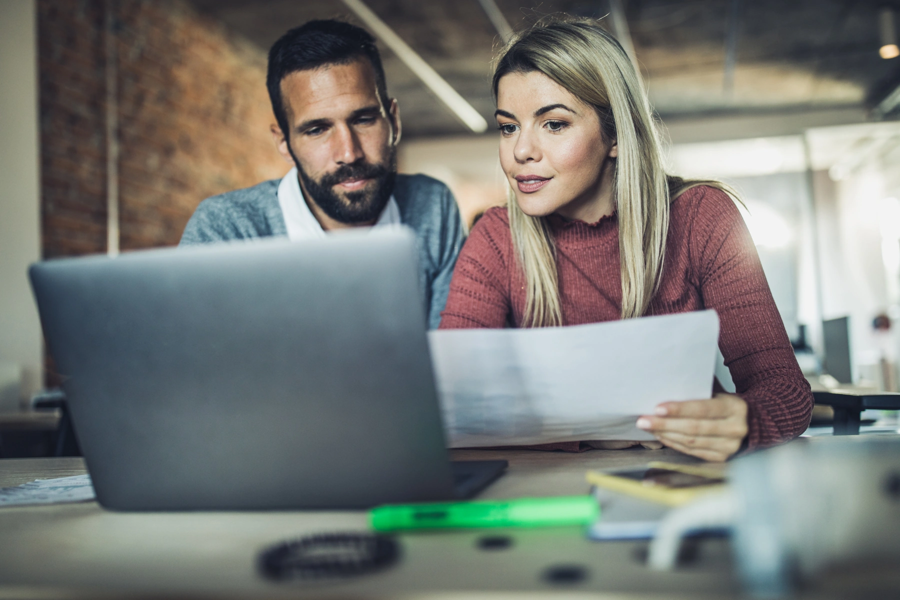 Twee collega’s bekijken samen documenten en een laptop op een kantoor.