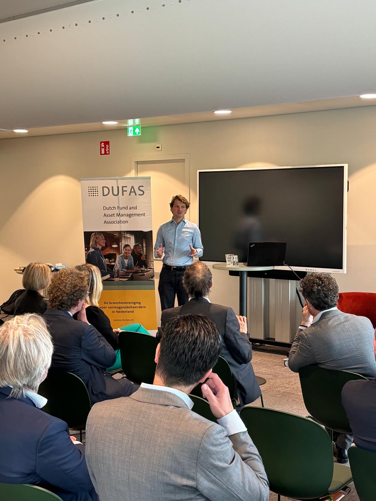 Een man geeft een presentatie voor een groep mensen bij een bijeenkomst van DUFAS, met een banner en scherm op de achtergrond.