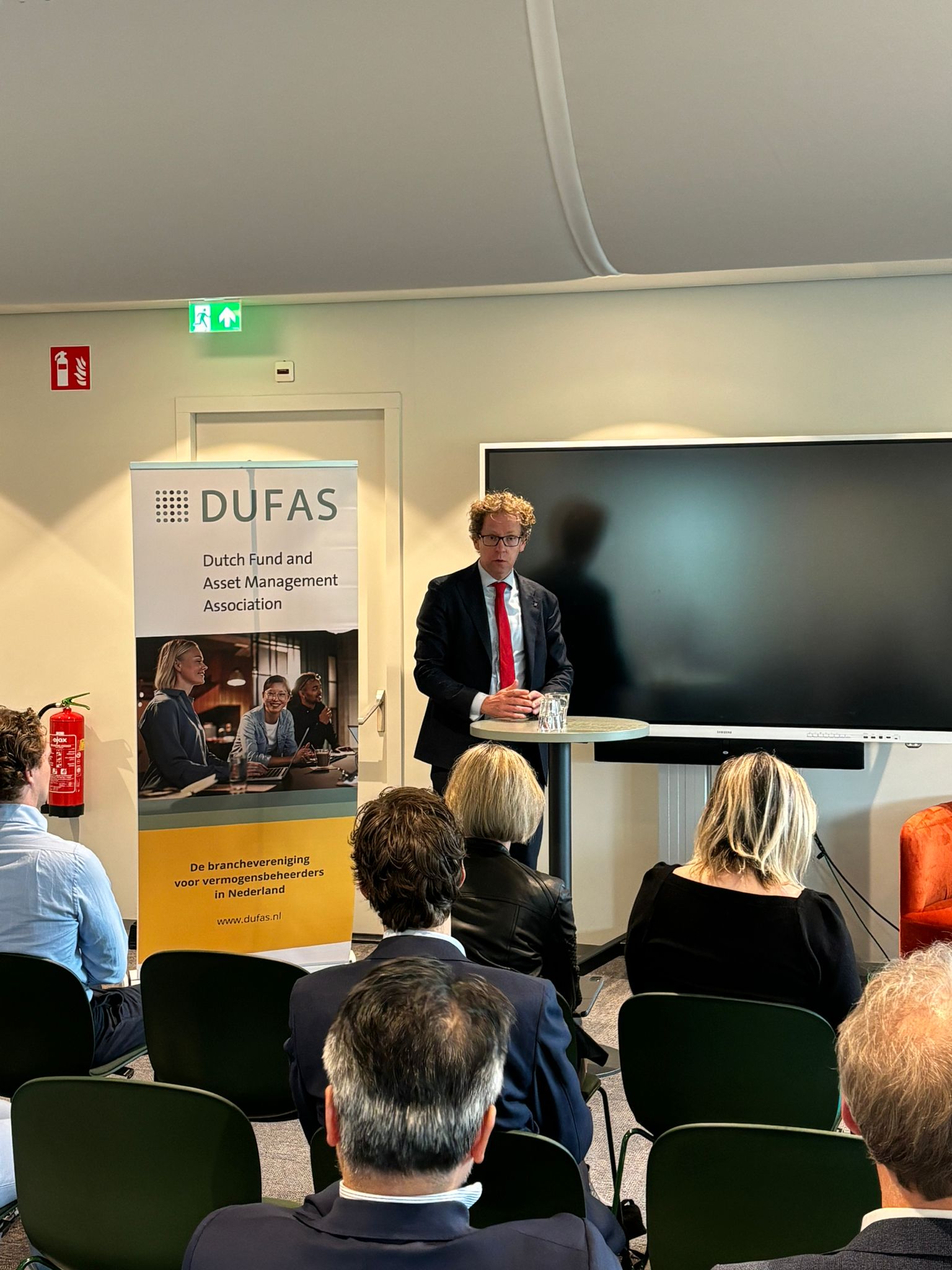 Man in pak spreekt voor publiek tijdens een DUFAS-bijeenkomst over vermogensbeheer.
