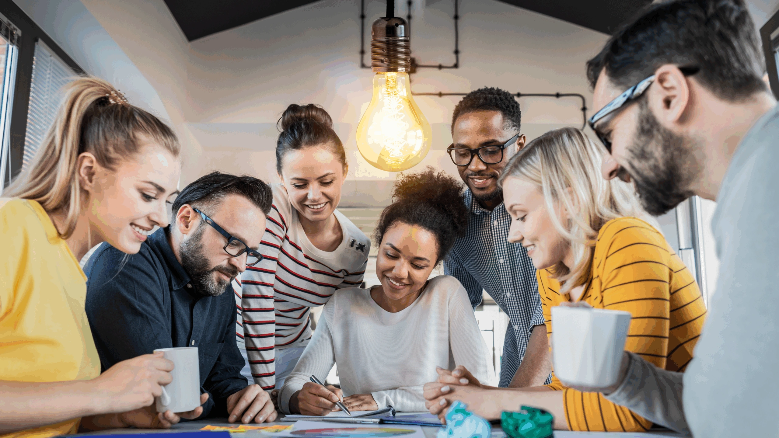 Een diverse groep jonge professionals werkt samen aan een creatieve brainstormsessie rond een tafel onder een brandende gloeilamp.