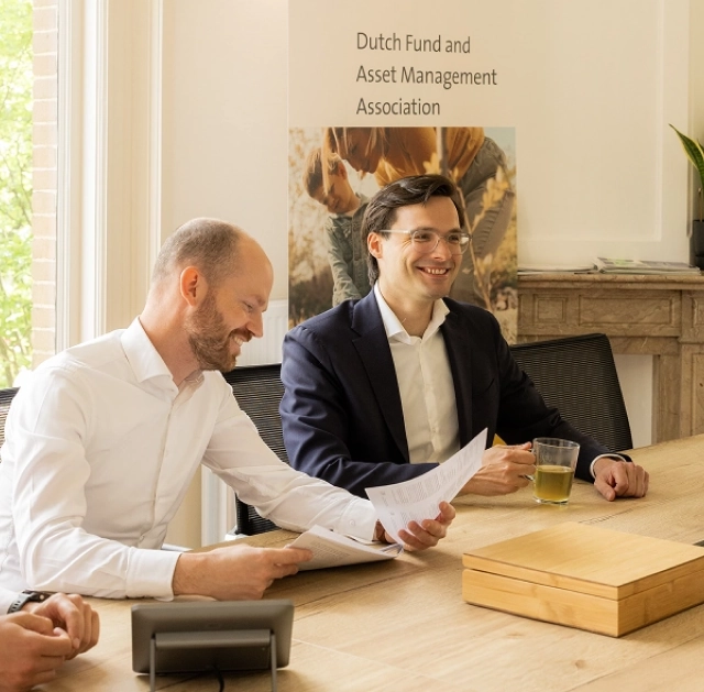 Twee mannen van het DUFAS-team zitten lachend aan een vergadertafel met documenten in de hand.