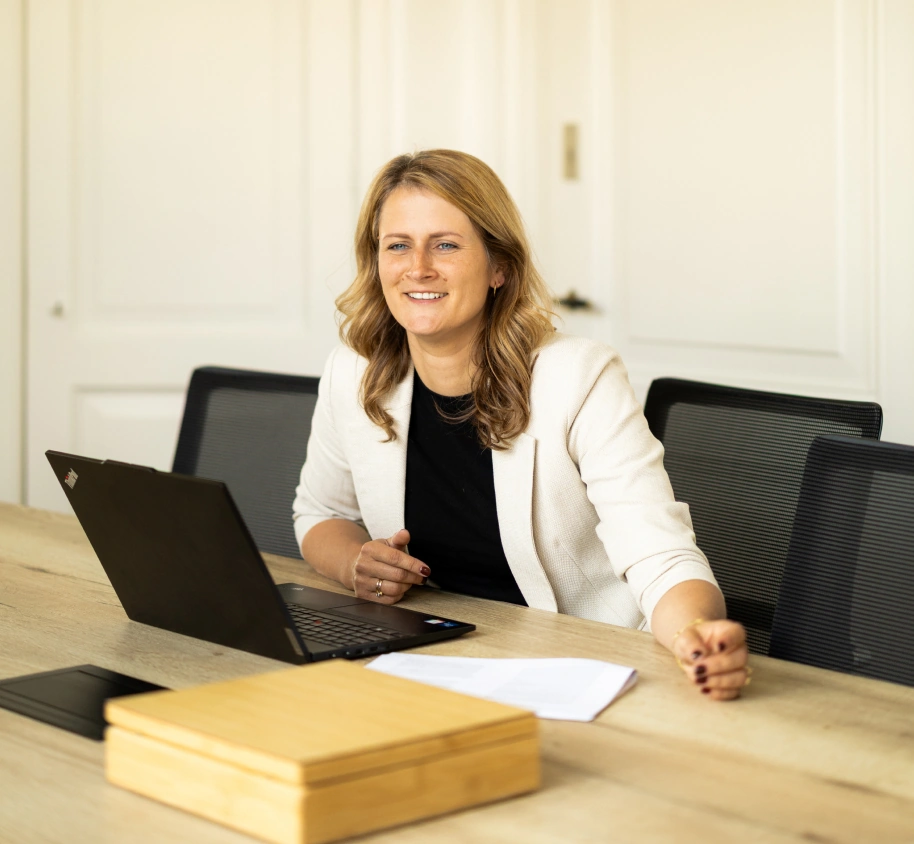 Vrouw genaamd Manouk zit glimlachend achter een laptop aan een vergadertafel.
