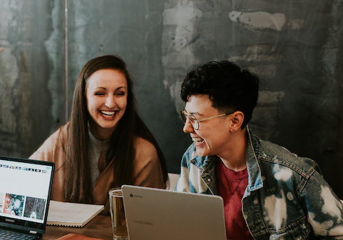 Twee DUFAS-collega's zitten lachend samen achter hun laptops in een informele werkomgeving.