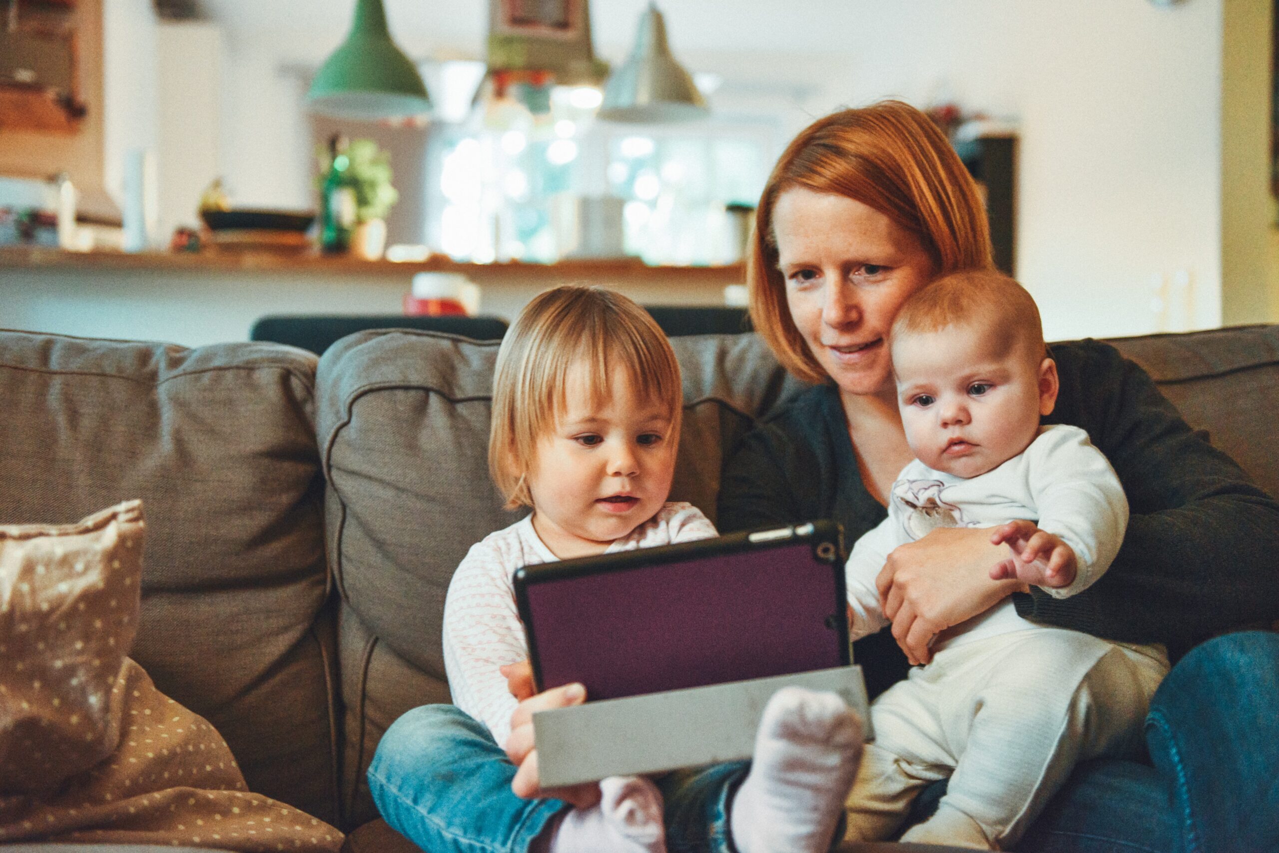Moeder zit met haar twee jonge kinderen op de bank en kijkt samen met hen naar een tablet.
