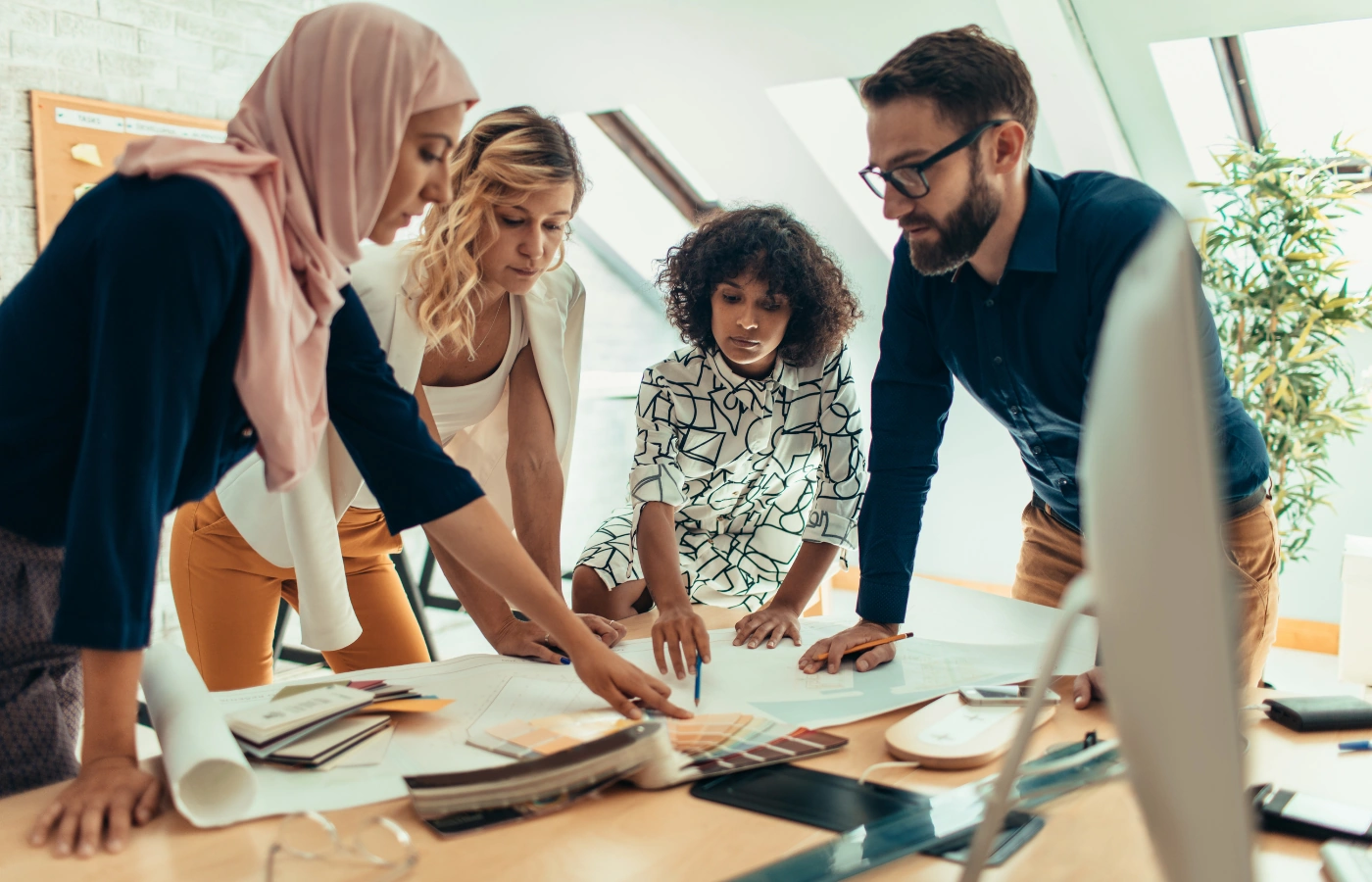 Een divers team van vier mensen werkt samen aan een project rond een tafel vol documenten en kleurstalen.